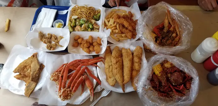 A table covered in orders: snow crab legs, fried fish, fried shrimp with crinkle fries, scallops and two boil bags